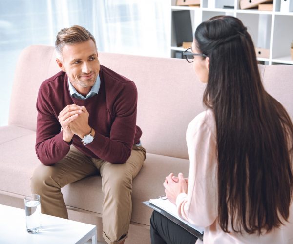 smiling patient sitting on couch and looking at psychotherapist with clipboard in office