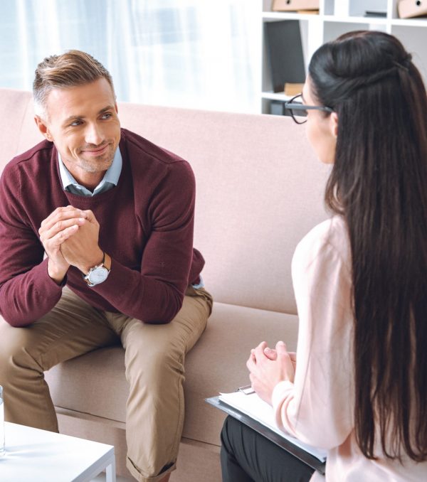 smiling patient sitting on couch and looking at psychotherapist with clipboard in office